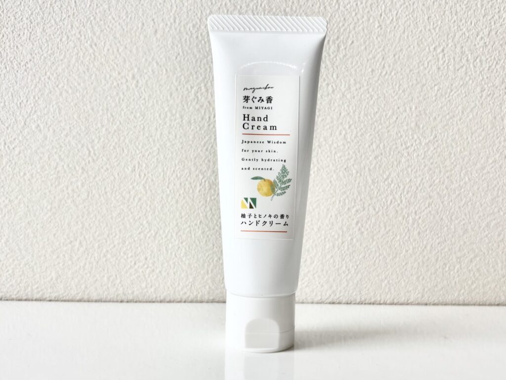 White squeeze tube of hand cream with a label featuring Japanese text and a citrus illustration, standing upright on a white surface against a textured wall.